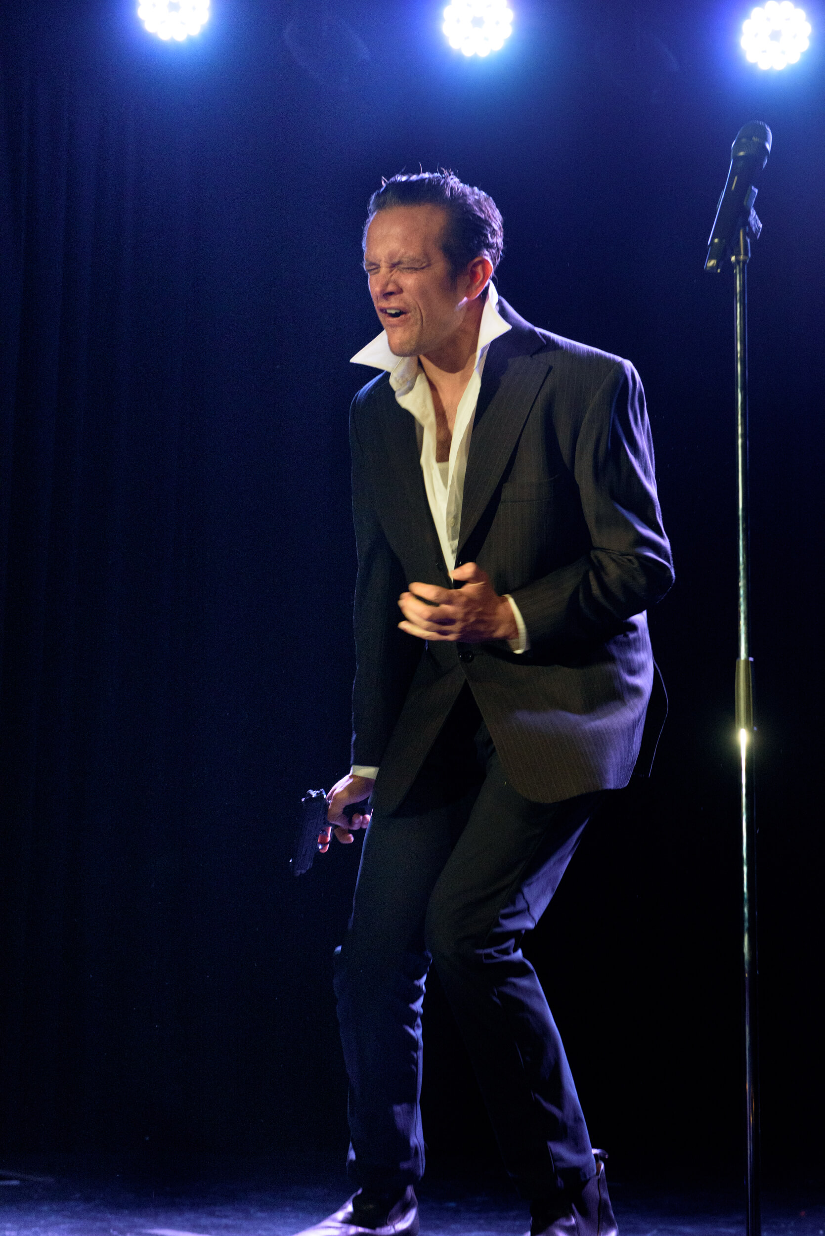 A performer in a black suit with a white shirt is holding a gun pointed at his foot. He has a pained expression on his face.