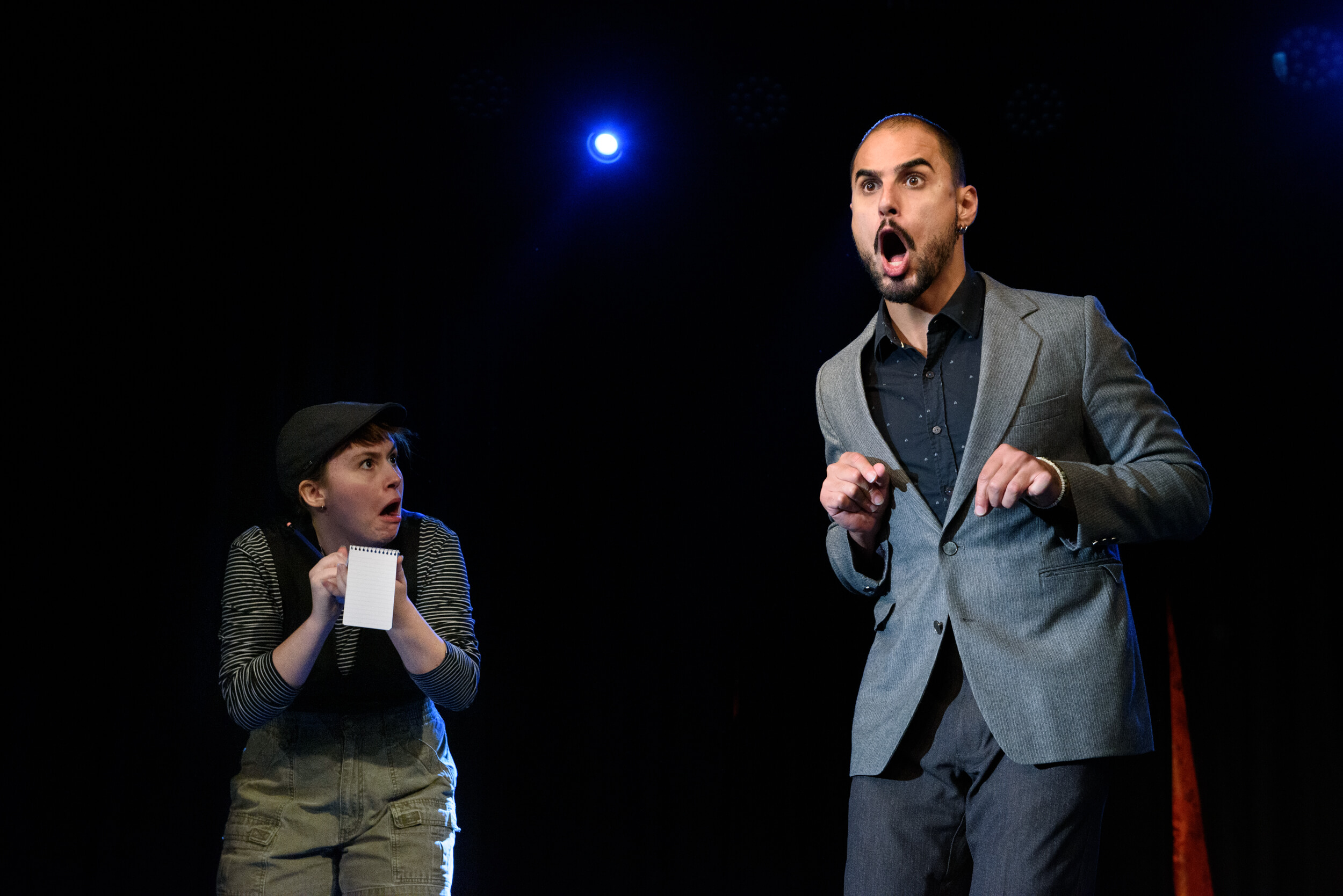 A performer wearing a grey suit is holding his hands up to his chest, imitating a lizard. Another performer is looking at him with a shocked expression. She is writing in a small notebook.