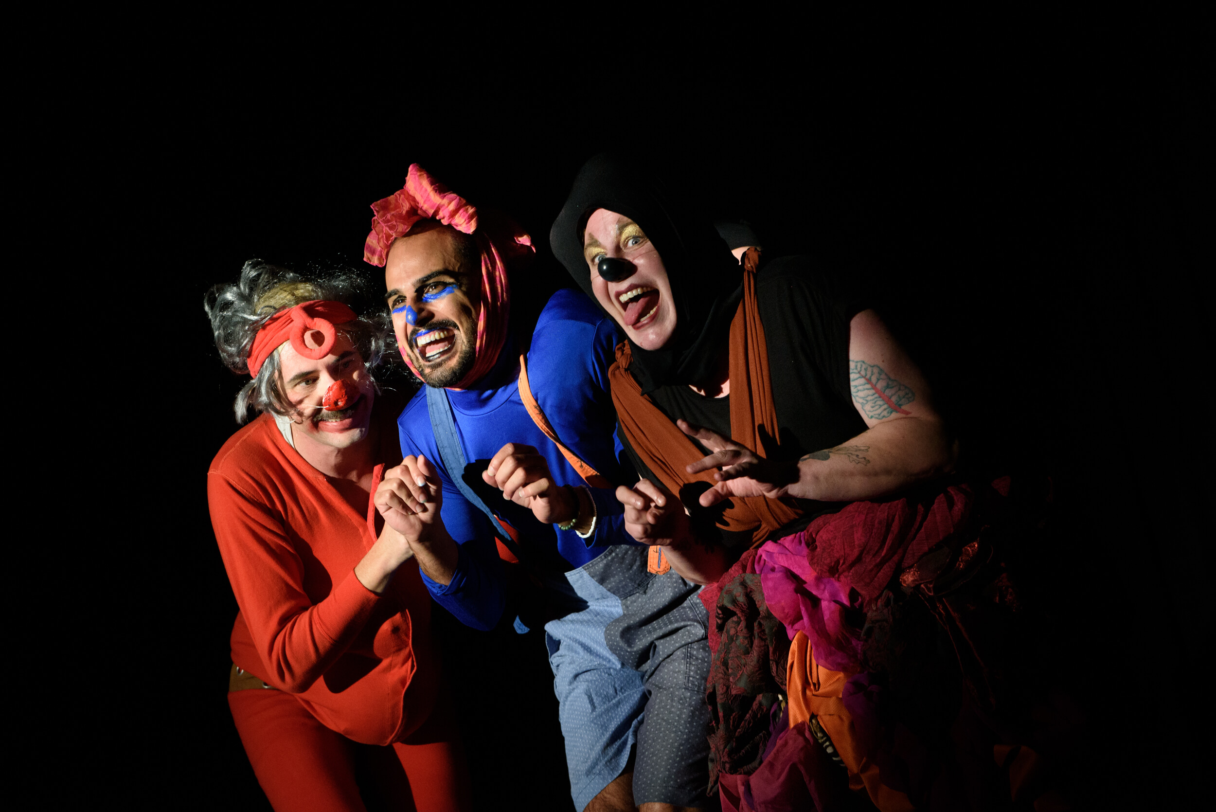 Three clowns in colourful costumes stand close together in a huddle. They are looking at the audience with mischievous expressions on their faces.