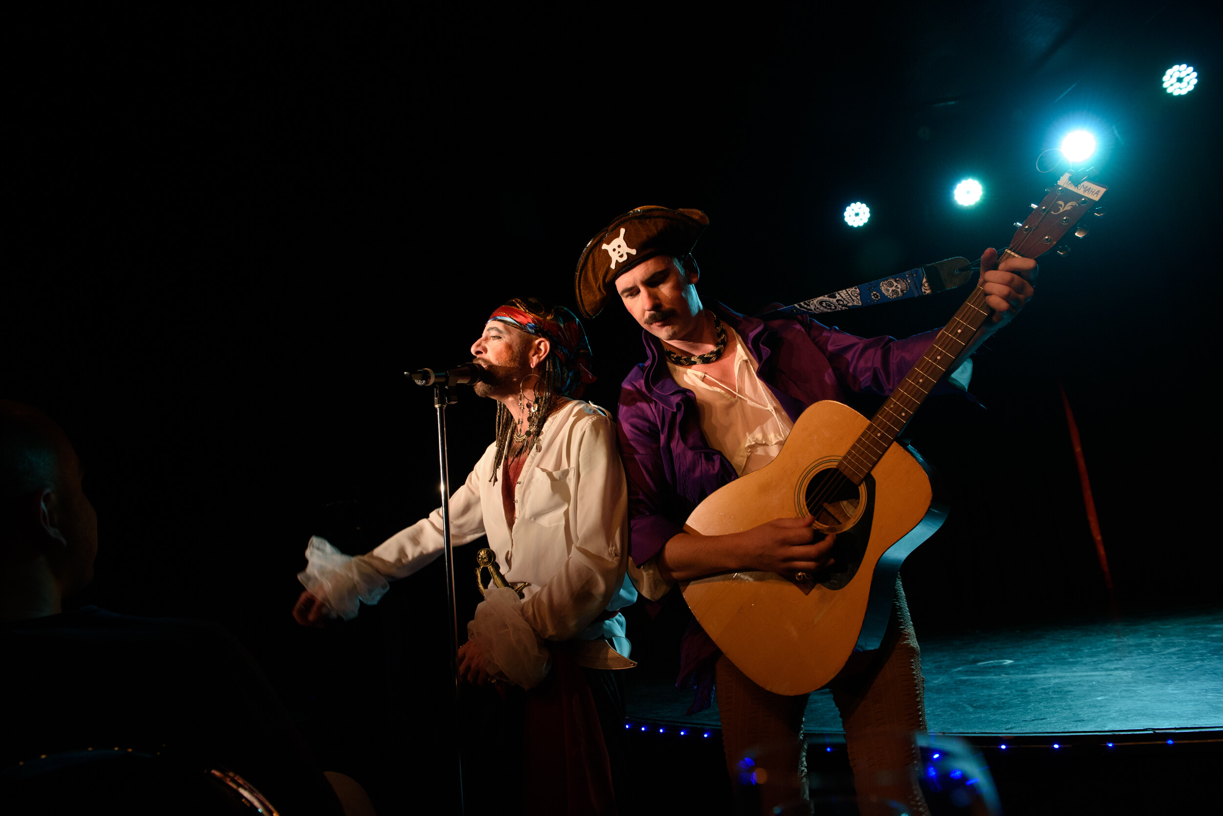 2 performers dressed as pirates stand shoulder to shoulder. One is singing into a microphone, the other is playing a guitar.